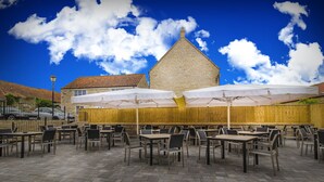 Un patio al aire libre con mesas y sillas, sombrillas y un edificio de piedra al fondo.
