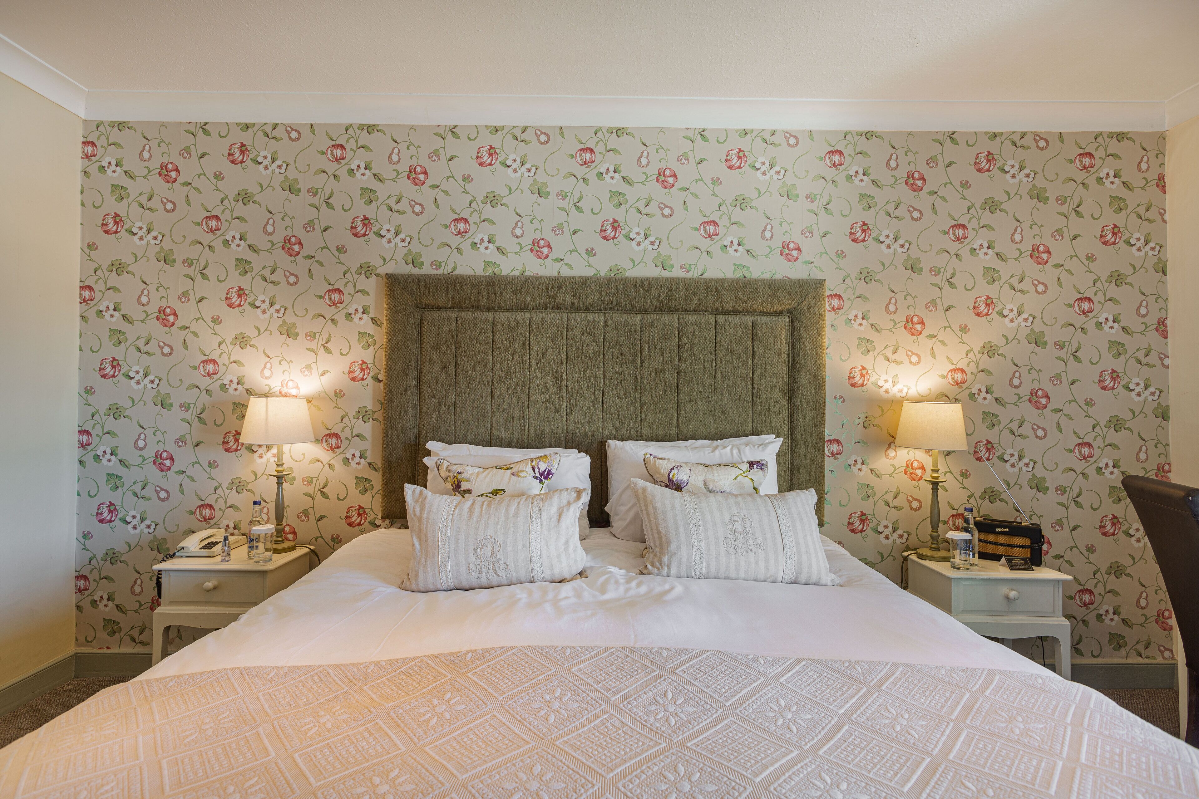 Una cama bien hecha con papel tapiz estampado con motivos florales, dos mesitas de noche con lámparas y un cabecero de madera.