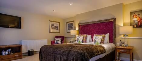 A well-lit bedroom with a large bed, a red armchair, a wooden nightstand, and a flat-screen TV mounted on the wall.