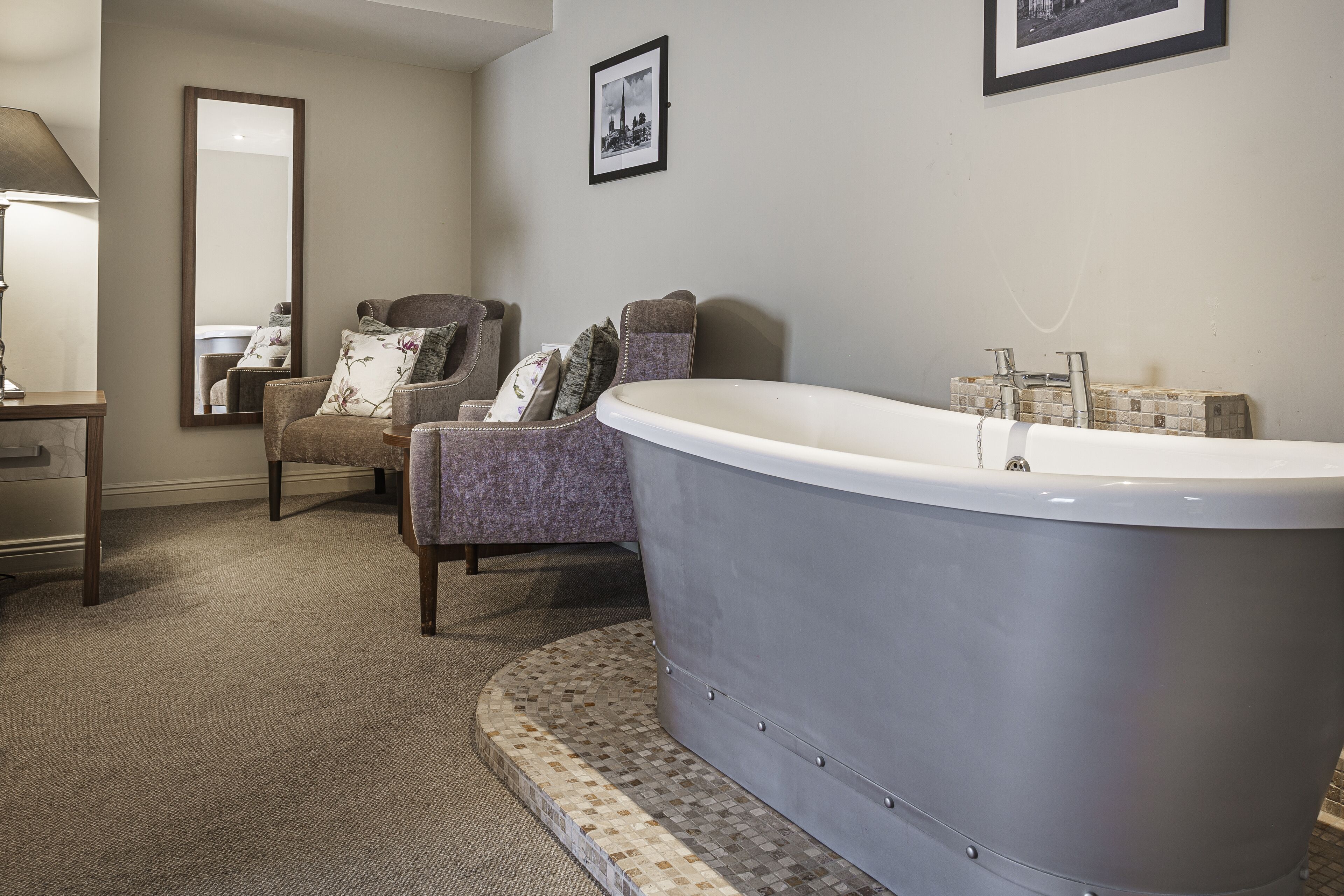 A bathroom with a freestanding bathtub, a chair with floral cushions, a mirror, and a framed picture on the wall.