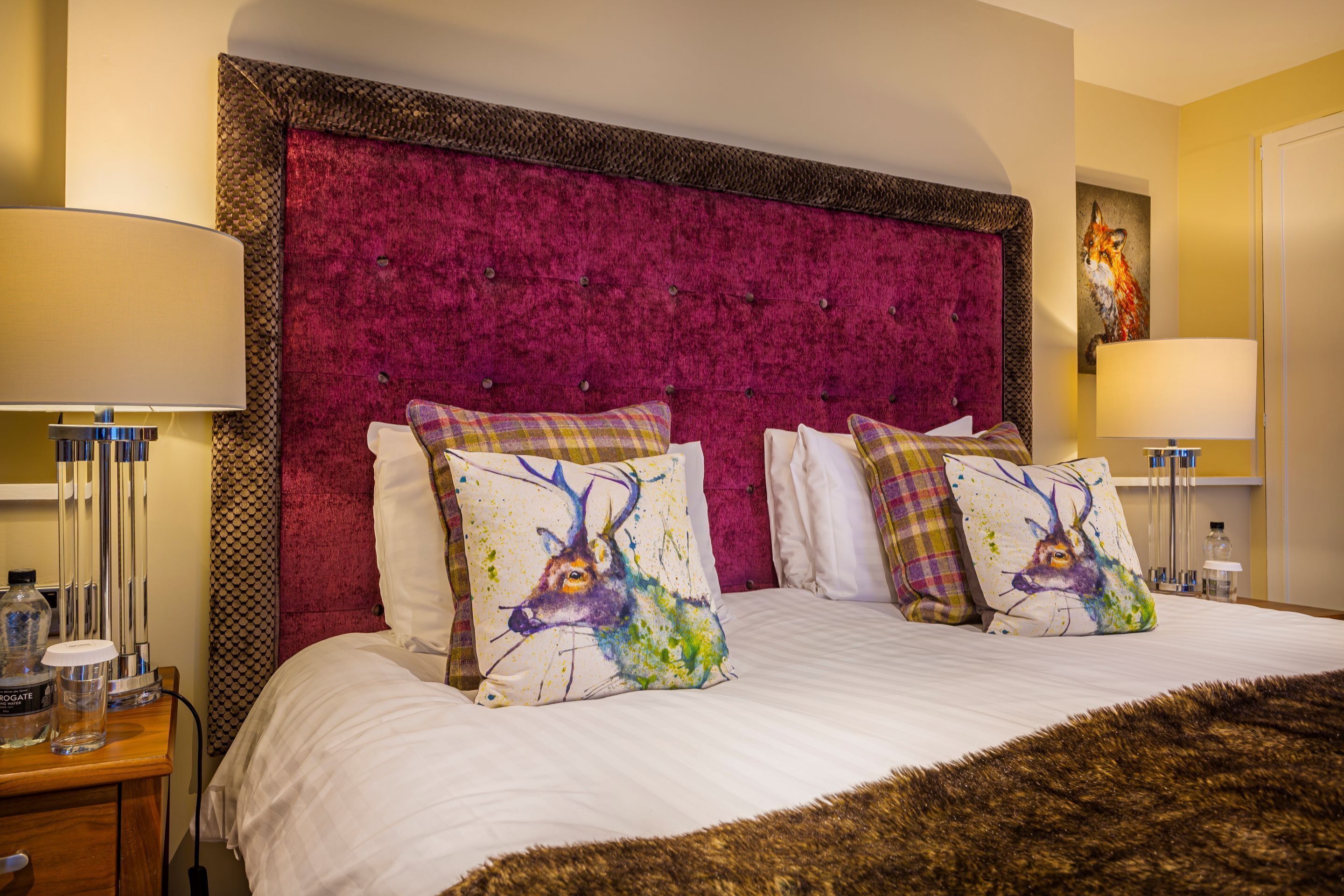 A hotel room with a large bed, two bedside lamps, a red upholstered headboard, and a painting of a fox on the wall.