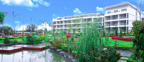 A modern hotel with a large garden, a pond, and a clear blue sky.