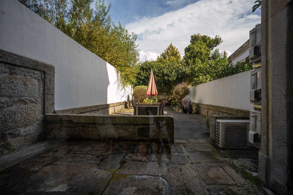 A courtyard with stone flooring, a table with chairs, and an umbrella. There are air conditioning units and a wall on the right.