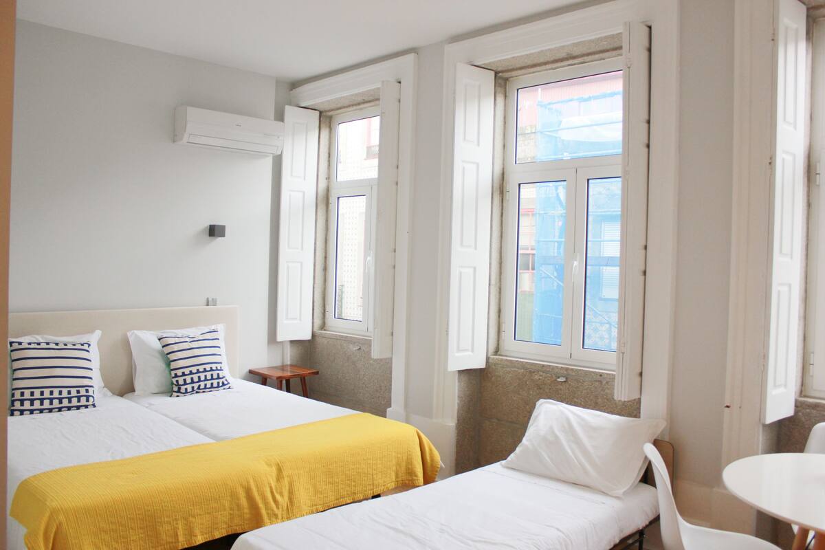 A hotel room with two beds, a small table, and a window with open shutters.