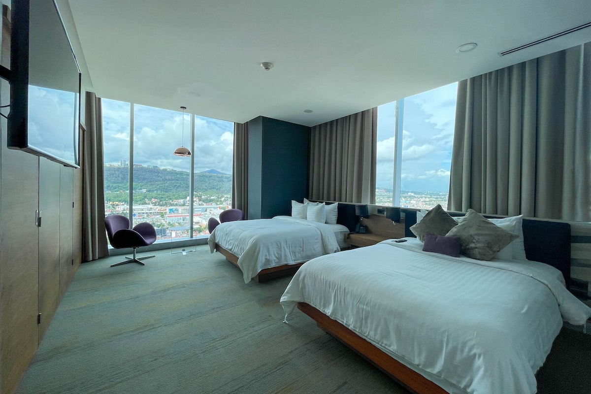 Habitación de hotel con dos camas, un ventanal grande con vista a la ciudad y una zona de estar con una silla y una mesa.