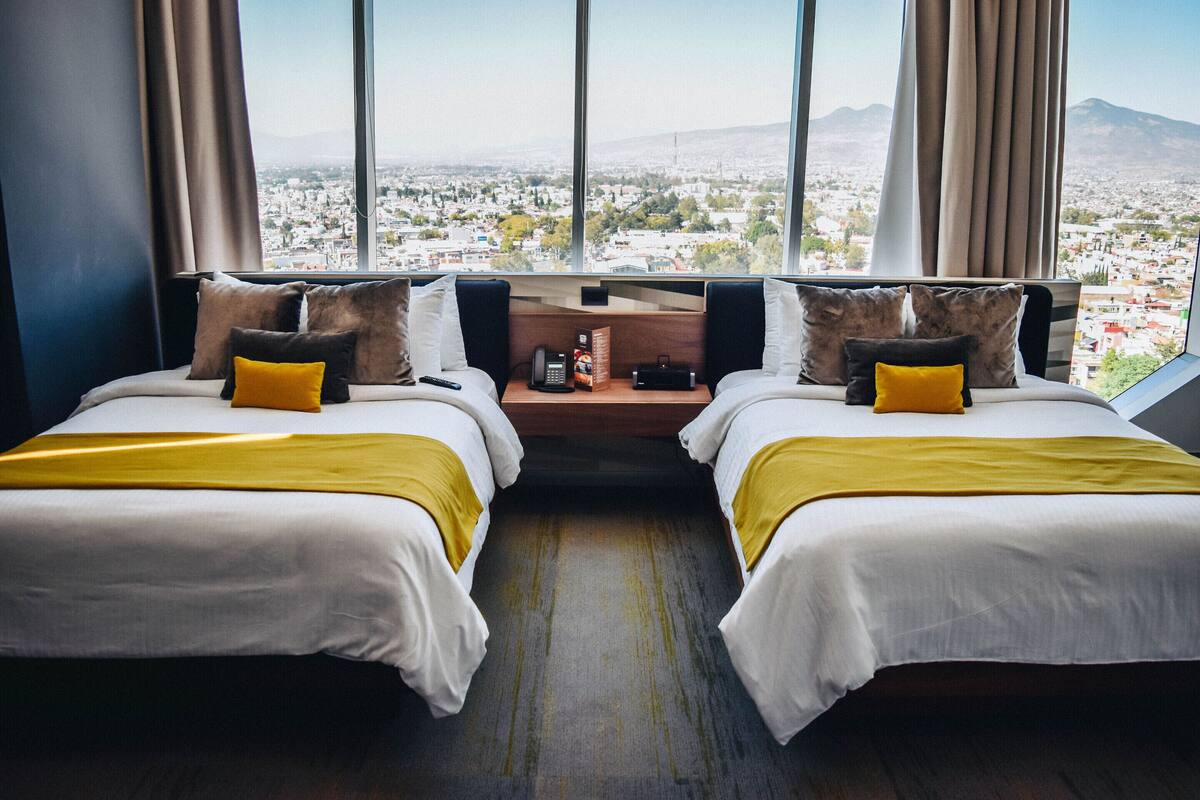 Habitación de hotel con dos camas, una mesita de noche de madera y vista a la ciudad a través de amplios ventanales.