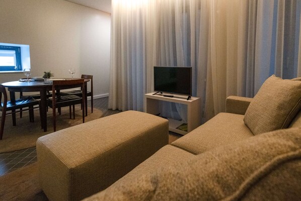 A living room with a sofa, ottoman, TV stand, and a dining area with a table and chairs. - Domum (Porto)
