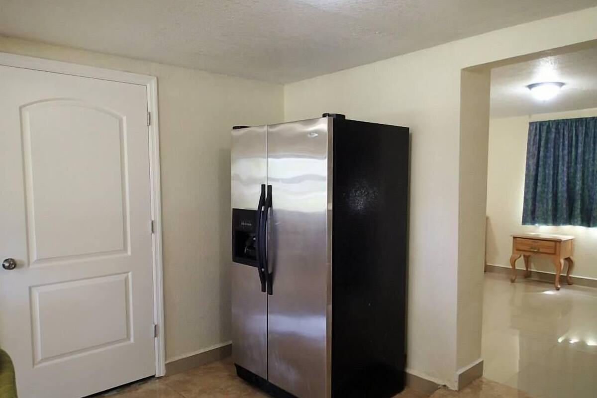 A compact kitchen with a stainless steel refrigerator, a white door, and a small wooden table with a lamp.
