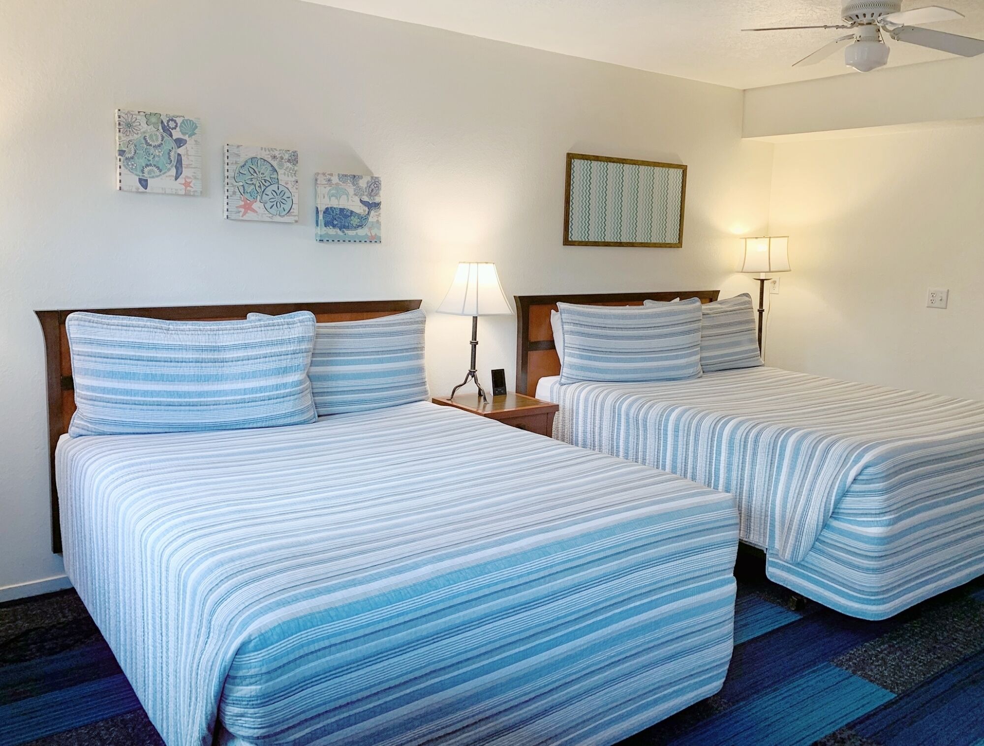 Ein Hotelzimmer mit zwei Betten, jeweils mit gestreifter Bettwäsche, einem Deckenventilator und Wandbildern.