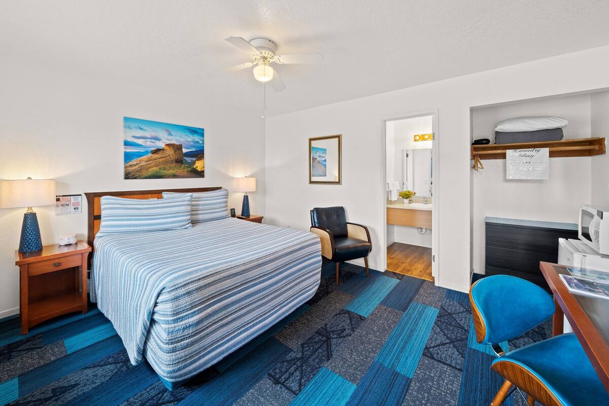 A hotel room with a bed, a desk, a chair, a ceiling fan, a lamp, a painting, and a laundry basket.