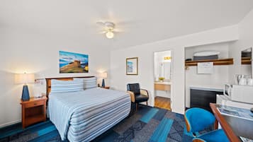 A hotel room with a bed, a desk, a chair, a ceiling fan, a painting, and a microwave.