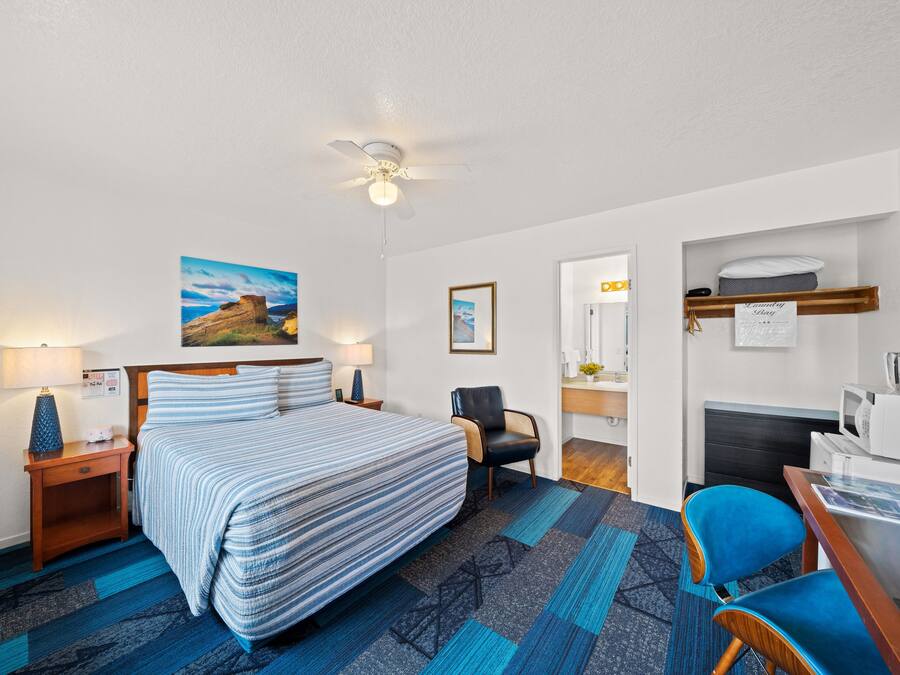 A hotel room with a bed, a desk, a chair, a ceiling fan, a painting, and a microwave.
