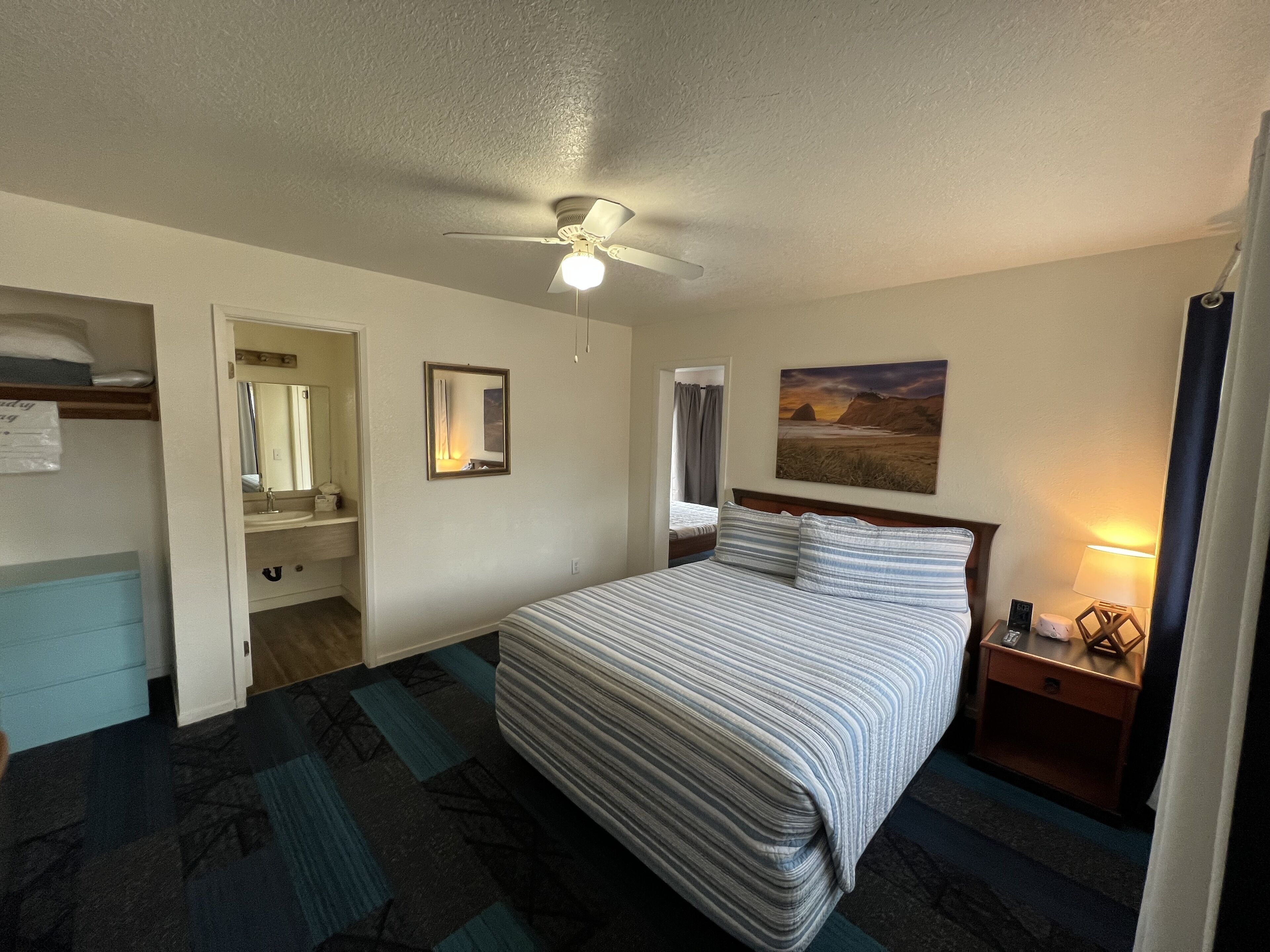 A hotel room with a bed, a nightstand, a lamp, a painting, a ceiling fan, and a bathroom visible through an open door.
