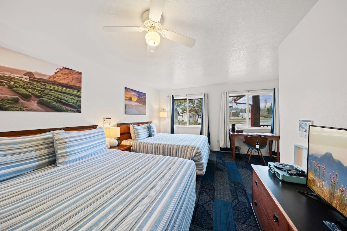 A hotel room with two beds, a desk, a chair, a ceiling fan, and a window with a view.