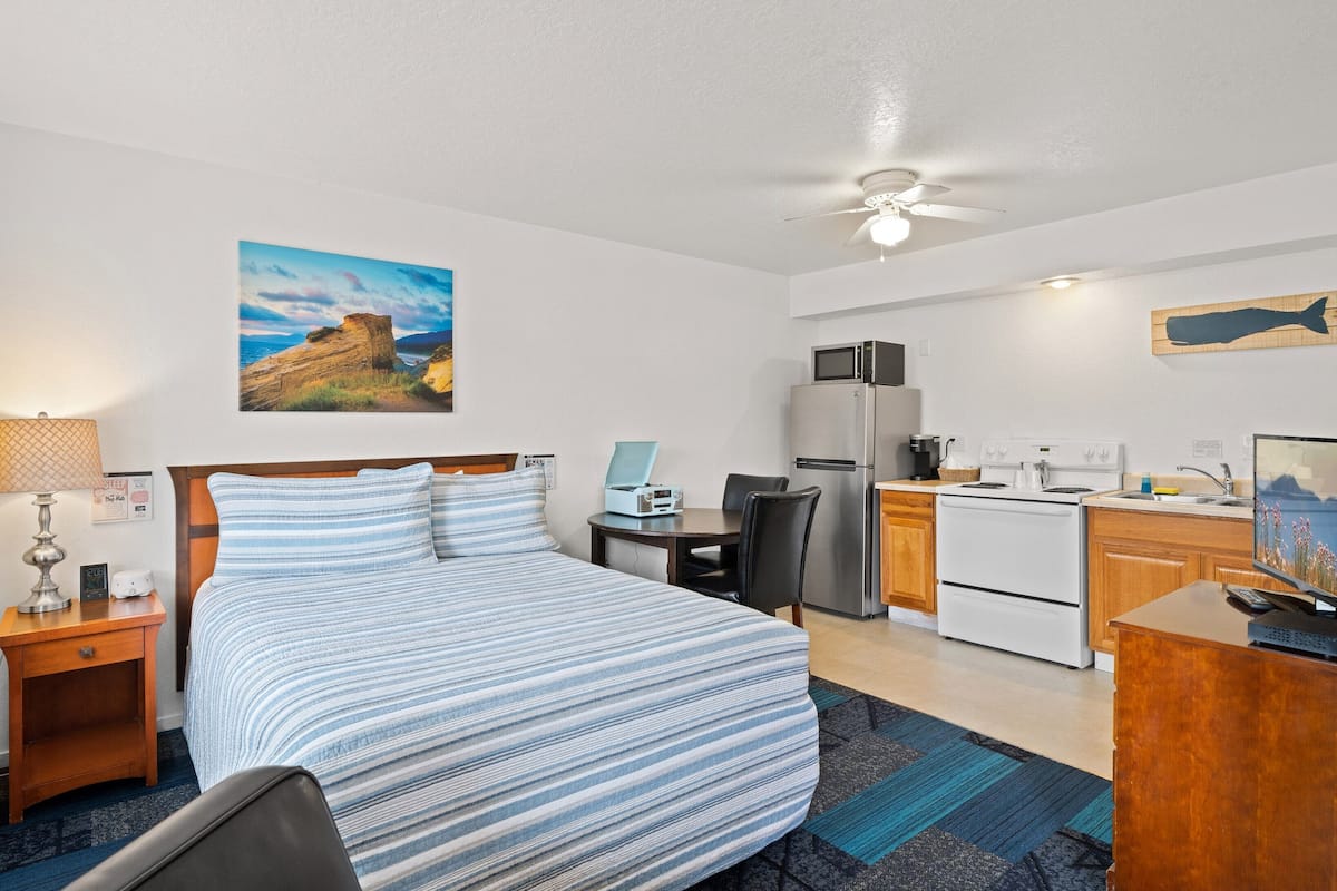 A hotel room with a bed, a desk, a kitchenette, and a ceiling fan.