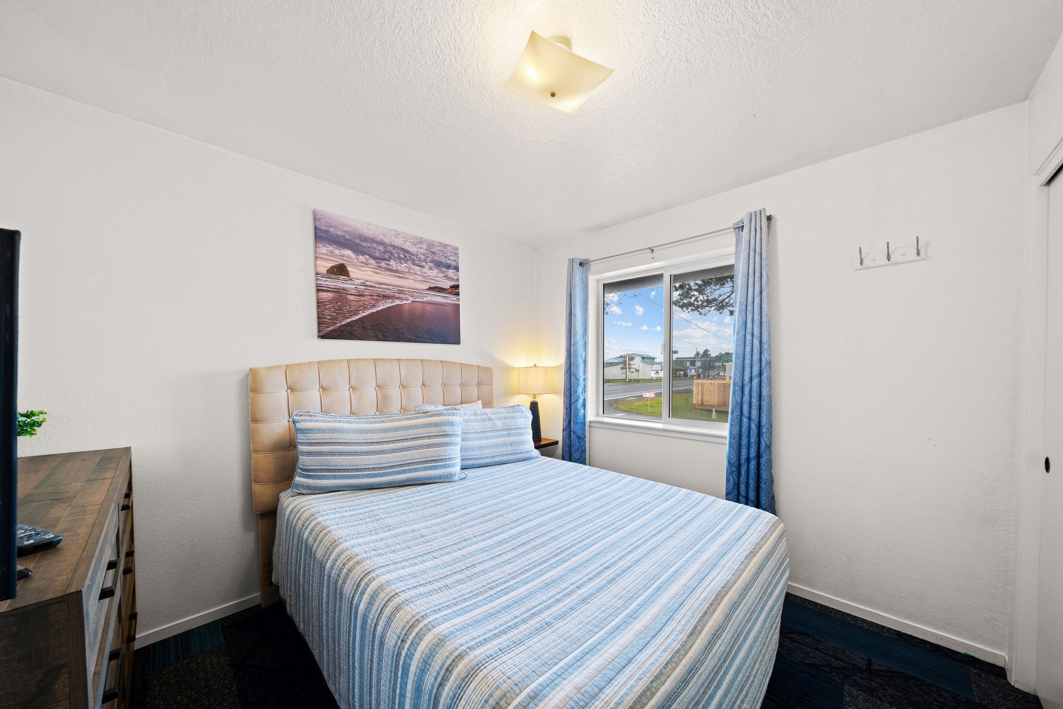A neatly arranged bedroom with a large bed, a headboard, a window with curtains, a wall-mounted lamp, and a painting.