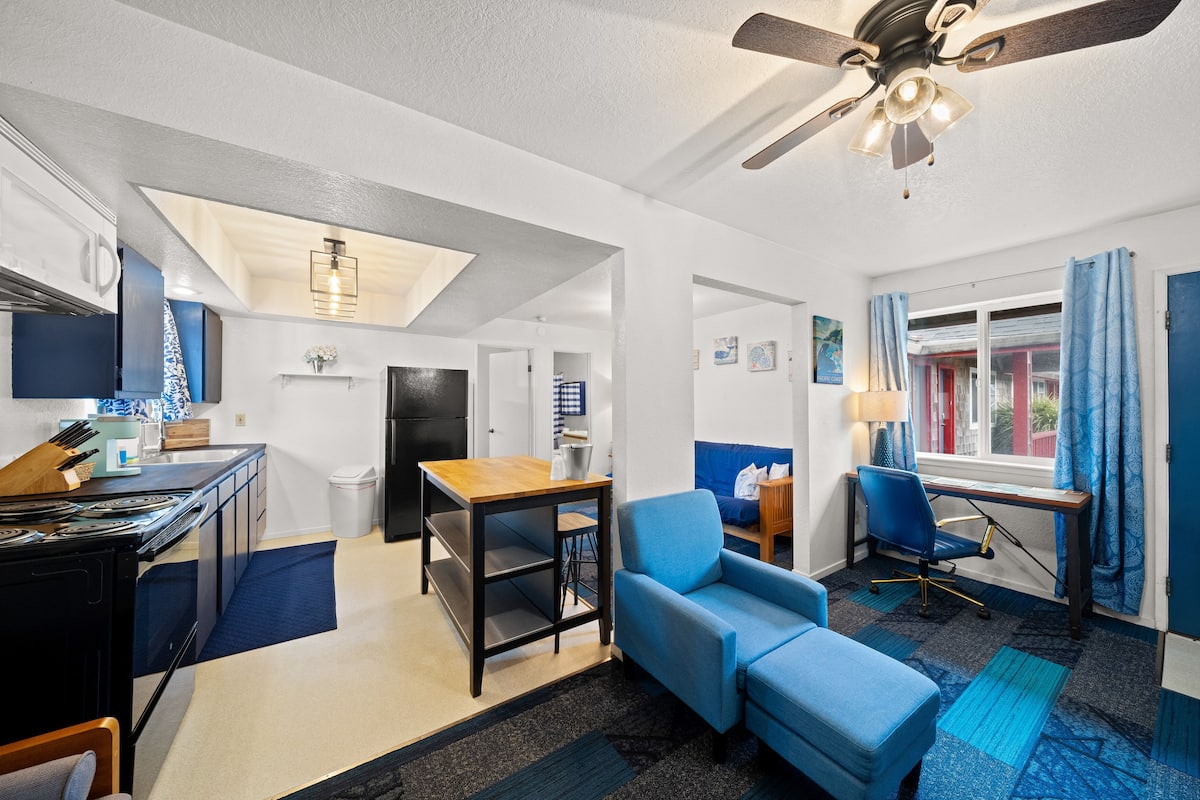 A compact living space with a kitchenette, a dining area, a blue sofa, a desk, and a ceiling fan.