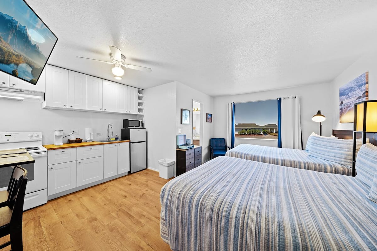 A hotel room with a kitchenette, two beds, a ceiling fan, and a view of the outdoors.