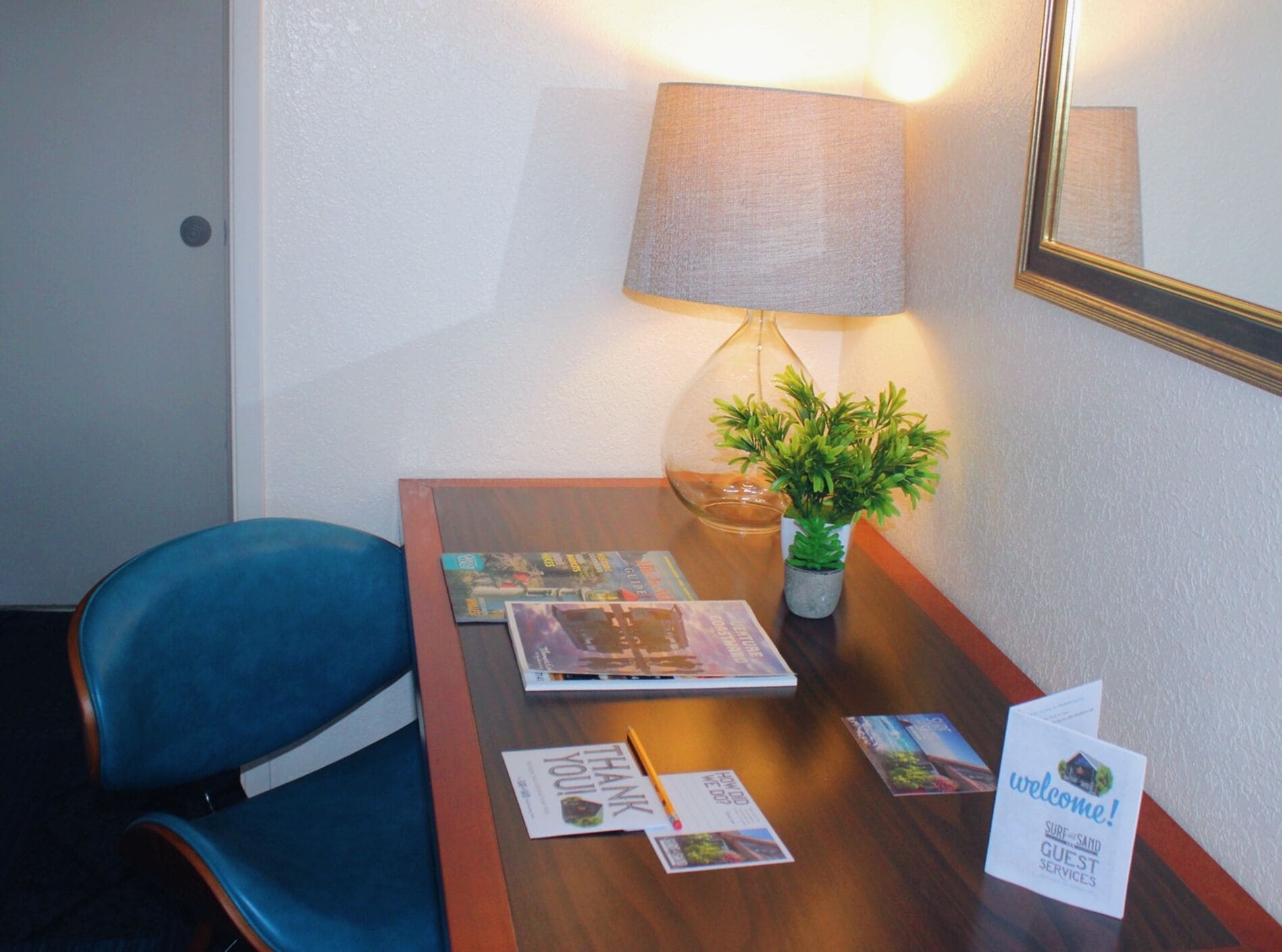A hotel room with a wooden desk, a blue chair, a lamp, a small potted plant, and a welcome sign.