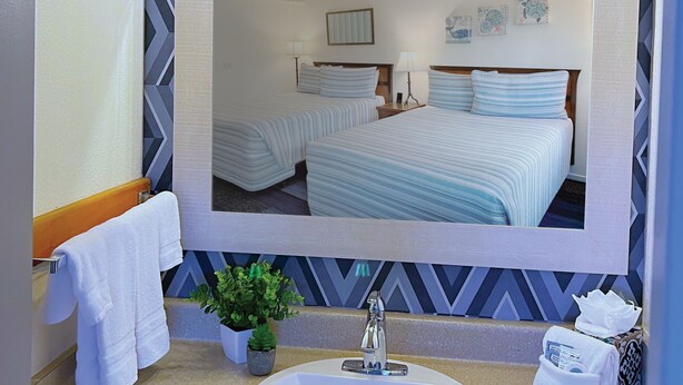 A bathroom with a large mirror, a sink, two towels, a potted plant, and a bed with blue bedding in the reflection. - Surf & Sand Inn (Pacific City)