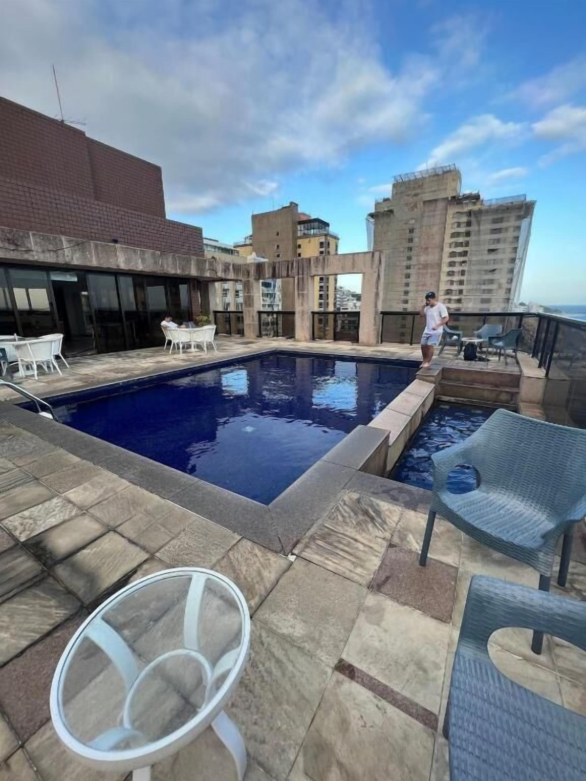 A rooftop pool area with a clear view of the city skyline, a seating area with chairs, and a person standing by the pool.
