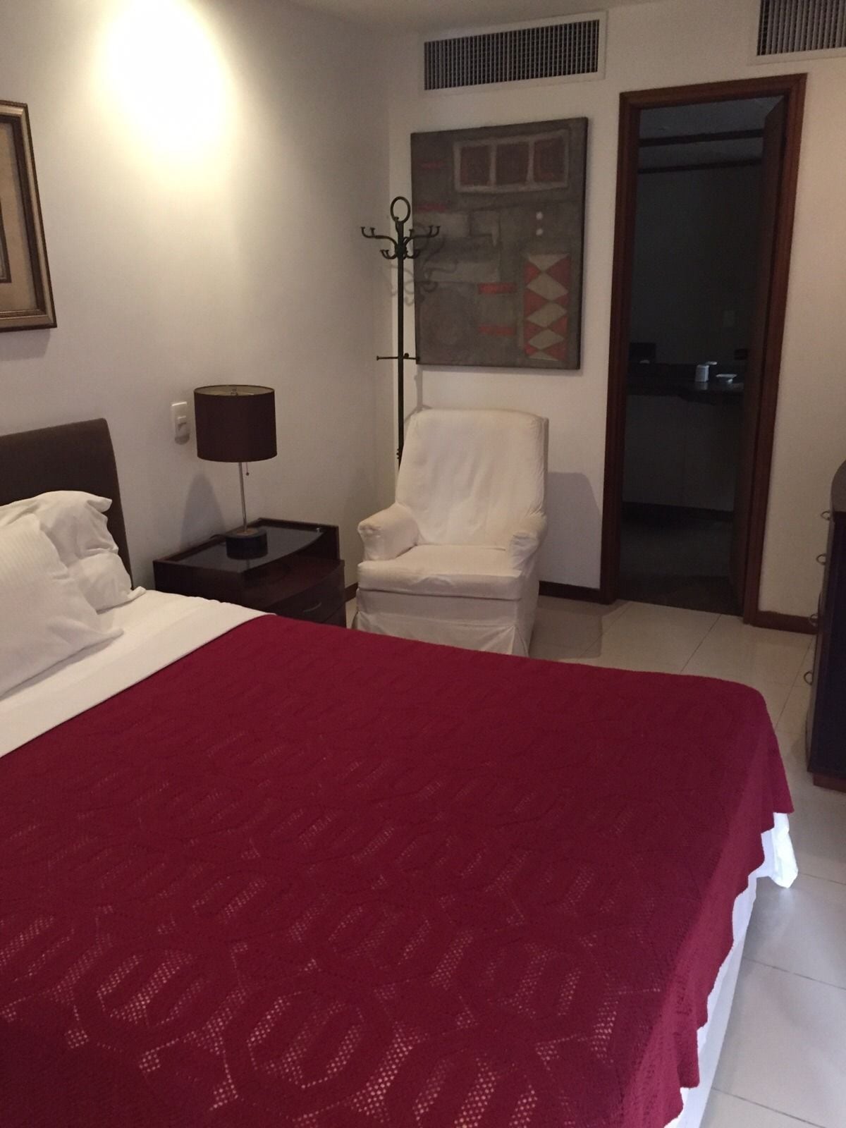 A hotel room with a large bed, a chair, a nightstand with a lamp, a painting on the wall, and a door leading to another room.