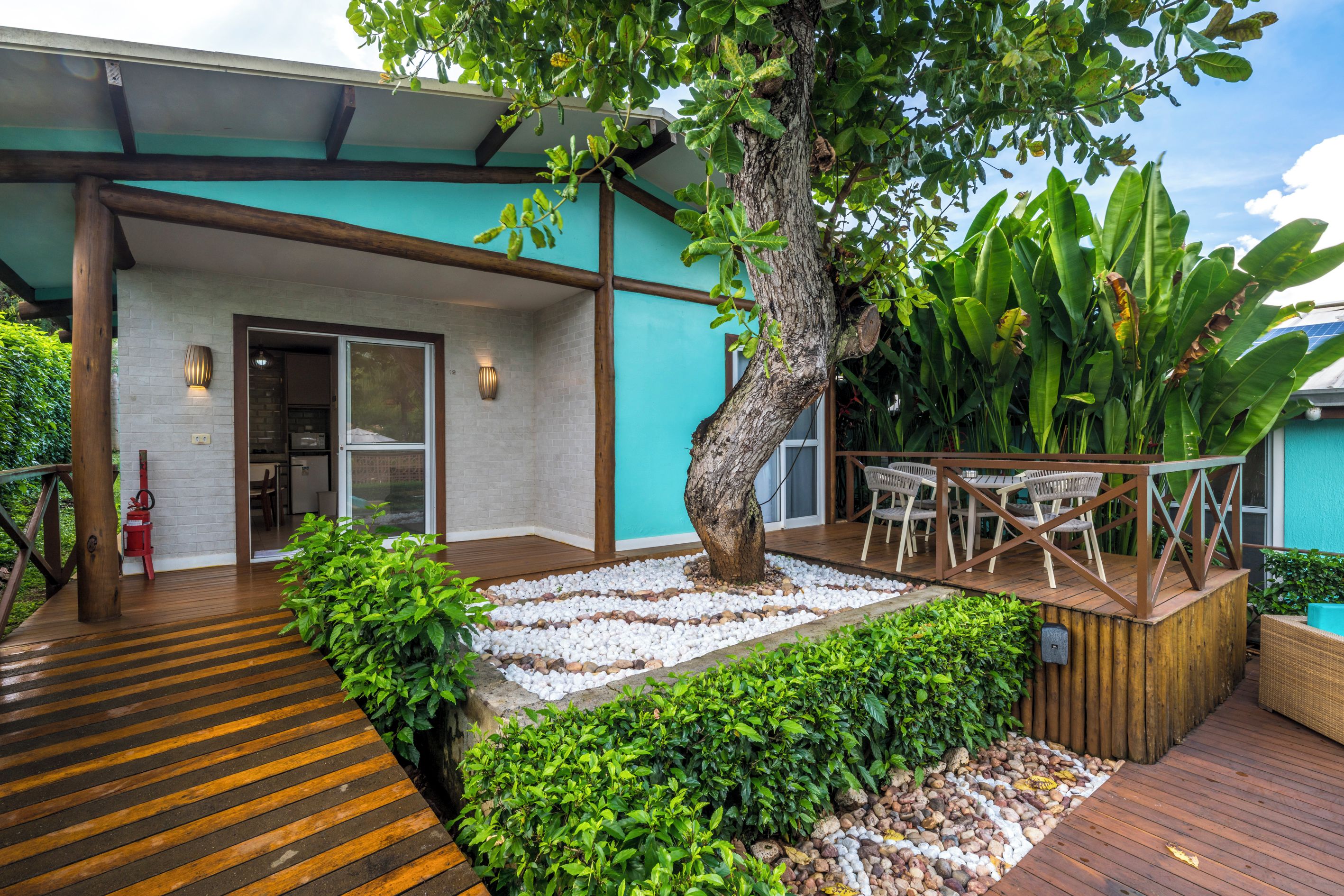 A covered outdoor area with a wooden deck, a seating area with chairs, and a tree.