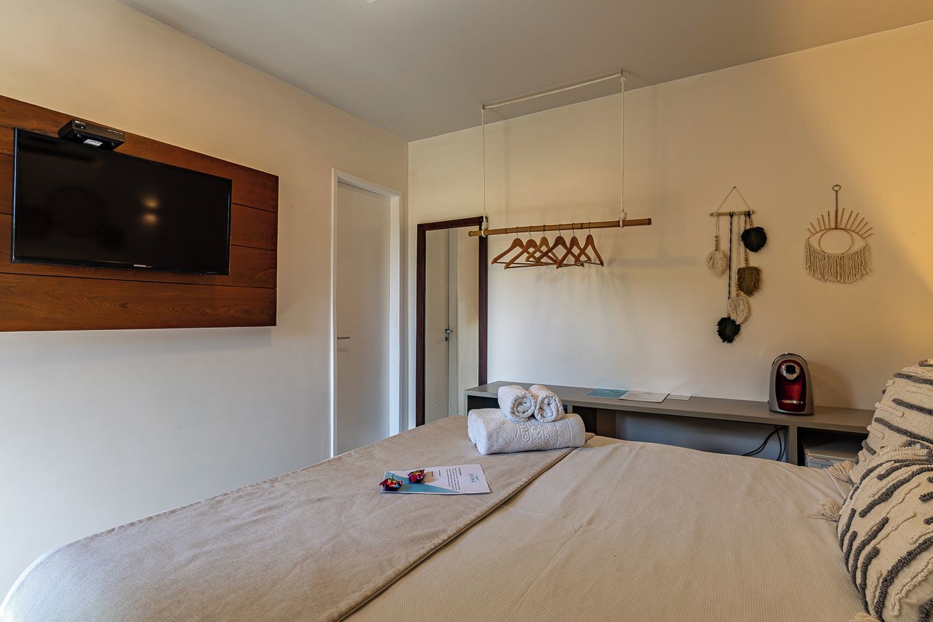 A hotel room with a bed, a flat-screen TV mounted on the wall, a bedside table with a kettle, and a wall-mounted towel rack.