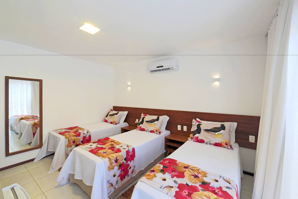A hotel room with three single beds, each with a floral bedspread and white sheets. There is a ceiling light, an air conditioning unit, and a mirror on the wall.