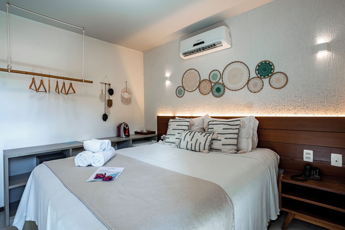 A hotel room with a bed, bedside tables, a wall-mounted air conditioner, decorative wall art, and a towel rack.