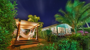 A well-lit outdoor area with a hammock, palm trees, and a building with large windows. - Pousada Atobá (Fernando de Noronha)