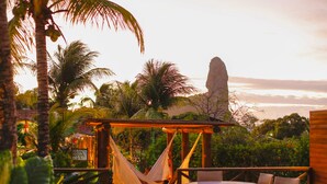A hammock under a wooden pergola, a round table with chairs, and a view of the ocean with a rock formation. - Pousada Atobá (Fernando de Noronha)