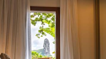 Ein Zimmer mit einem Bett, einem kleinen Tisch mit einem gefalteten Handtuch und Blick auf eine natürliche Landschaft durch ein Fenster mit Vorhängen.