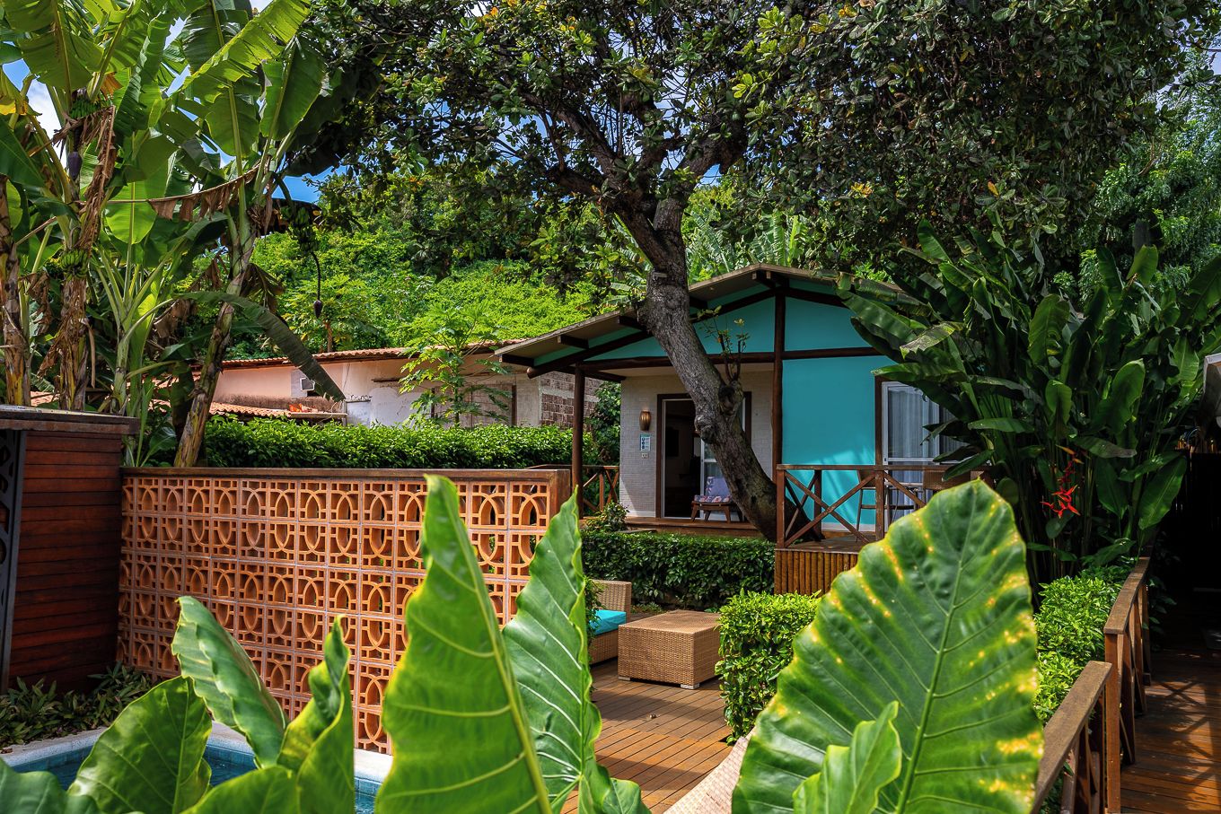 A tropical garden with a wooden deck, a covered seating area, and a building with a green roof.