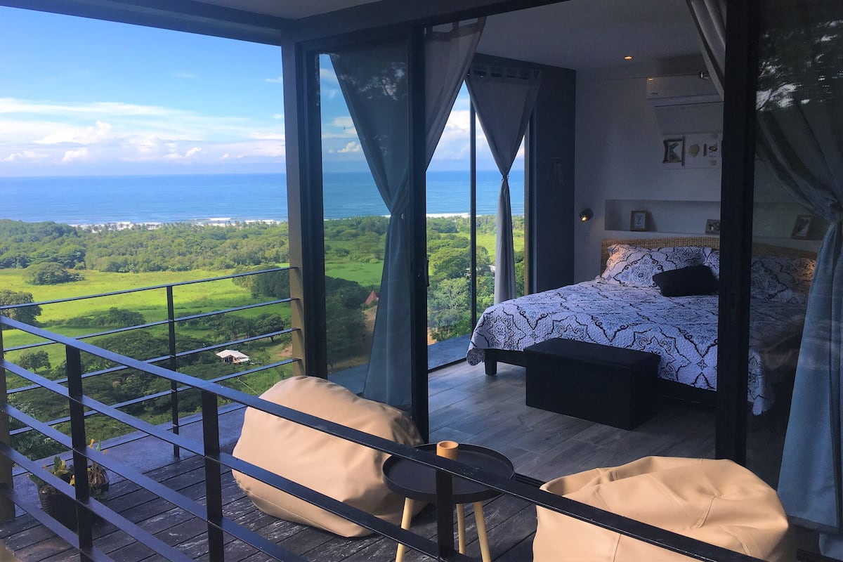 Een slaapkamer met een groot raam met uitzicht op zee en groen.