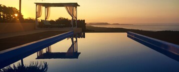Una piscina infinita tranquila con cubierta de madera y un toldo que da a una playa al atardecer.