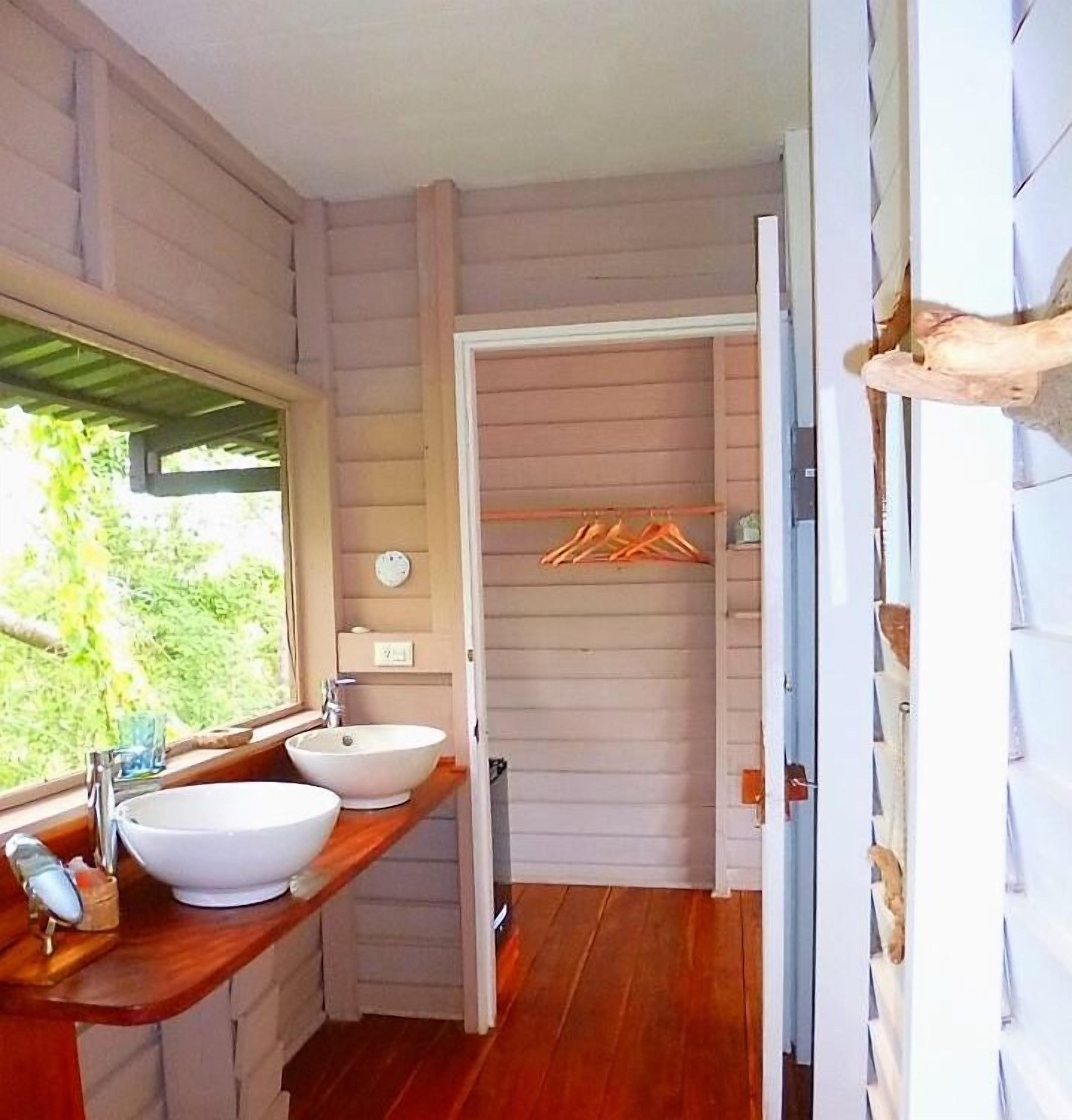 Baño con dos lavamanos, encimeras de madera, una ventana y una puerta que da a un armario con ropa colgada.