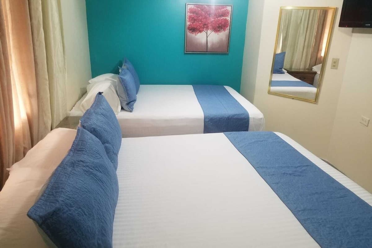 A hotel room with two beds, each with blue bedding and pillows. There is a painting of a tree on the wall, a mirror, and a television mounted on the wall.