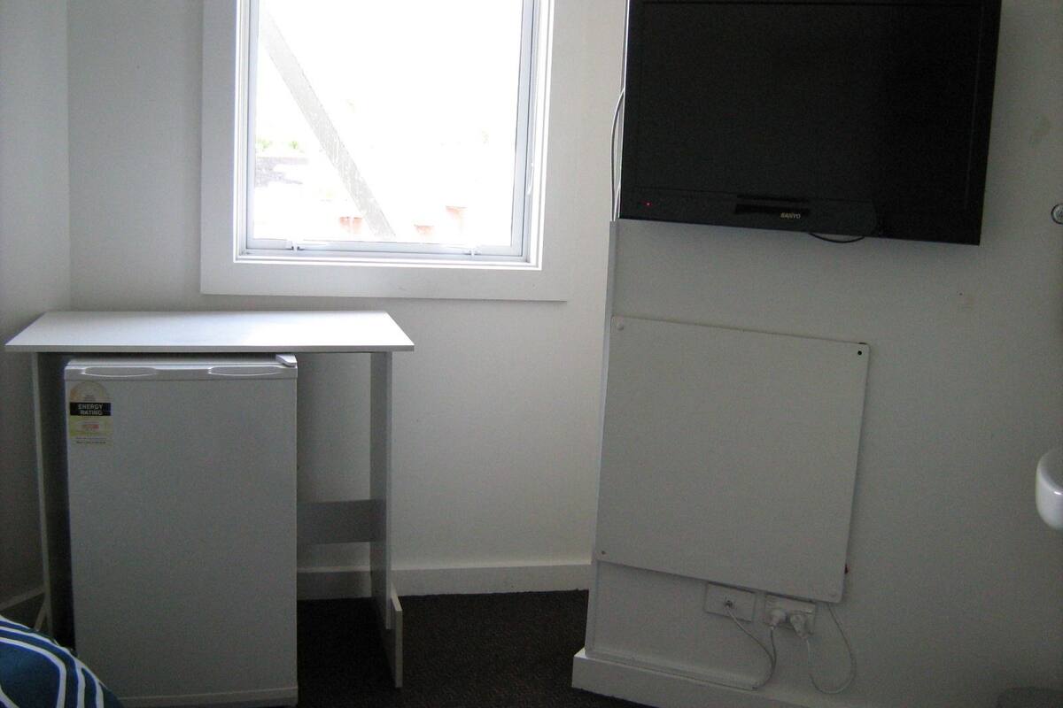 A small room with a white desk, a white refrigerator, a television mounted on the wall, and a window providing natural light.