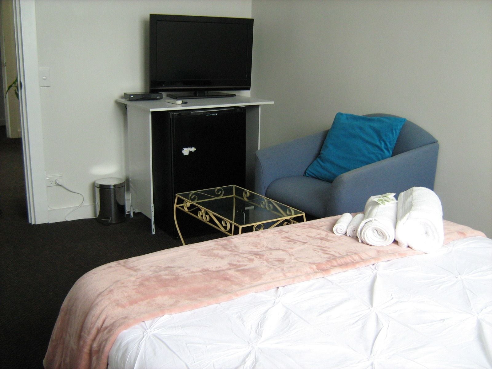 Habitación de hotel con cama, televisor sobre un pedestal, una mesita, silla con cojín y un cesto de basura.