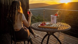 A woman sitting on a chair on a wooden deck with a table in front of her. The table has a bottle of champagne, a glass, and a tray with food. - Alkantmooi Nature Reserve Keurkloof Units (Plettenberg Bay)