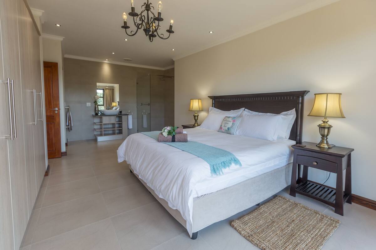 A bedroom with a large bed, bedside lamps, a chandelier, and a bathroom in the background.