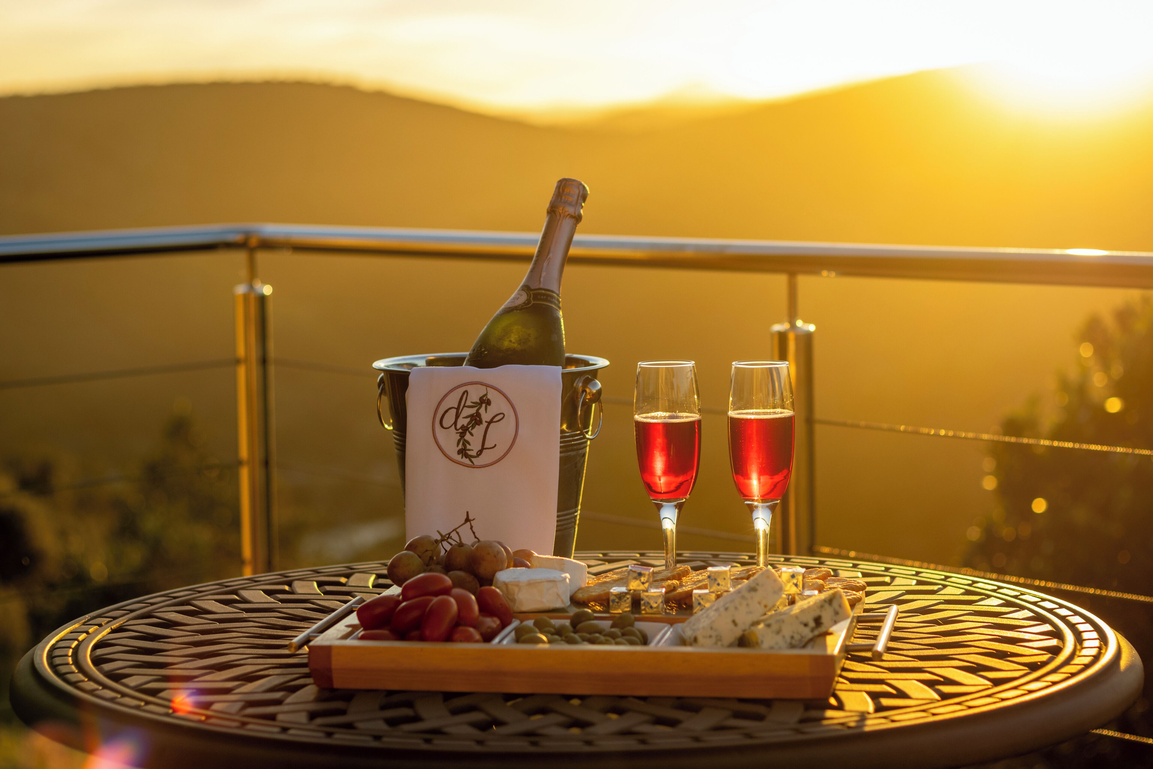 Une table avec une bouteille de champagne, deux verres, des raisins, du fromage et des crackers, le tout placé sur un balcon d’où l’on peut admirer le coucher de soleil.