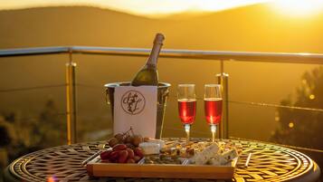 Une table avec une bouteille de champagne, deux verres, des raisins, du fromage et des crackers, le tout placé sur un balcon d’où l’on peut admirer le coucher de soleil.