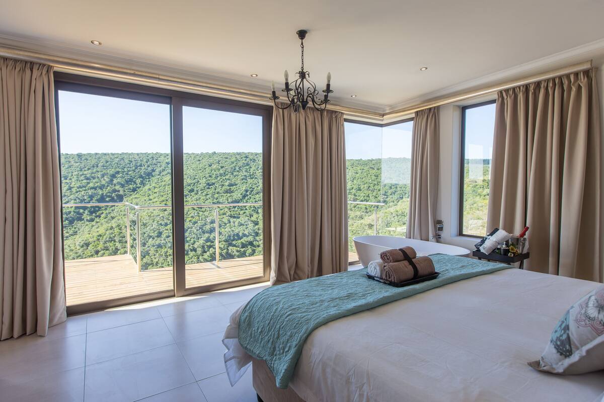 A bedroom with a large bed, a chandelier, a view of a forested landscape, and a balcony.