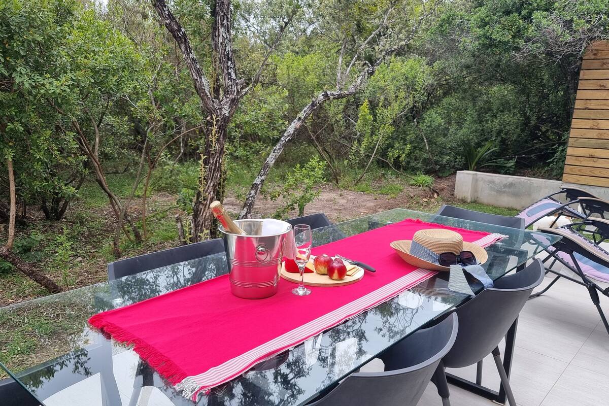 An outdoor dining area with a glass table, metal chairs, and a red tablecloth. There is a bottle of champagne, a glass, and a plate with food and a hat on the table.