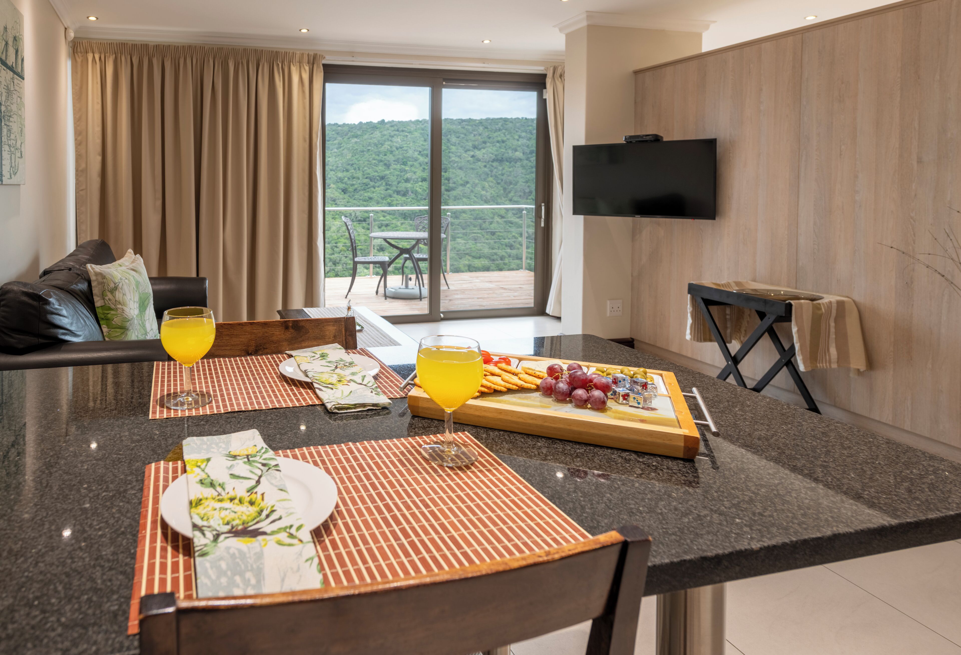A modern dining area with a table set for two, a TV mounted on the wall, and a balcony with a scenic view.