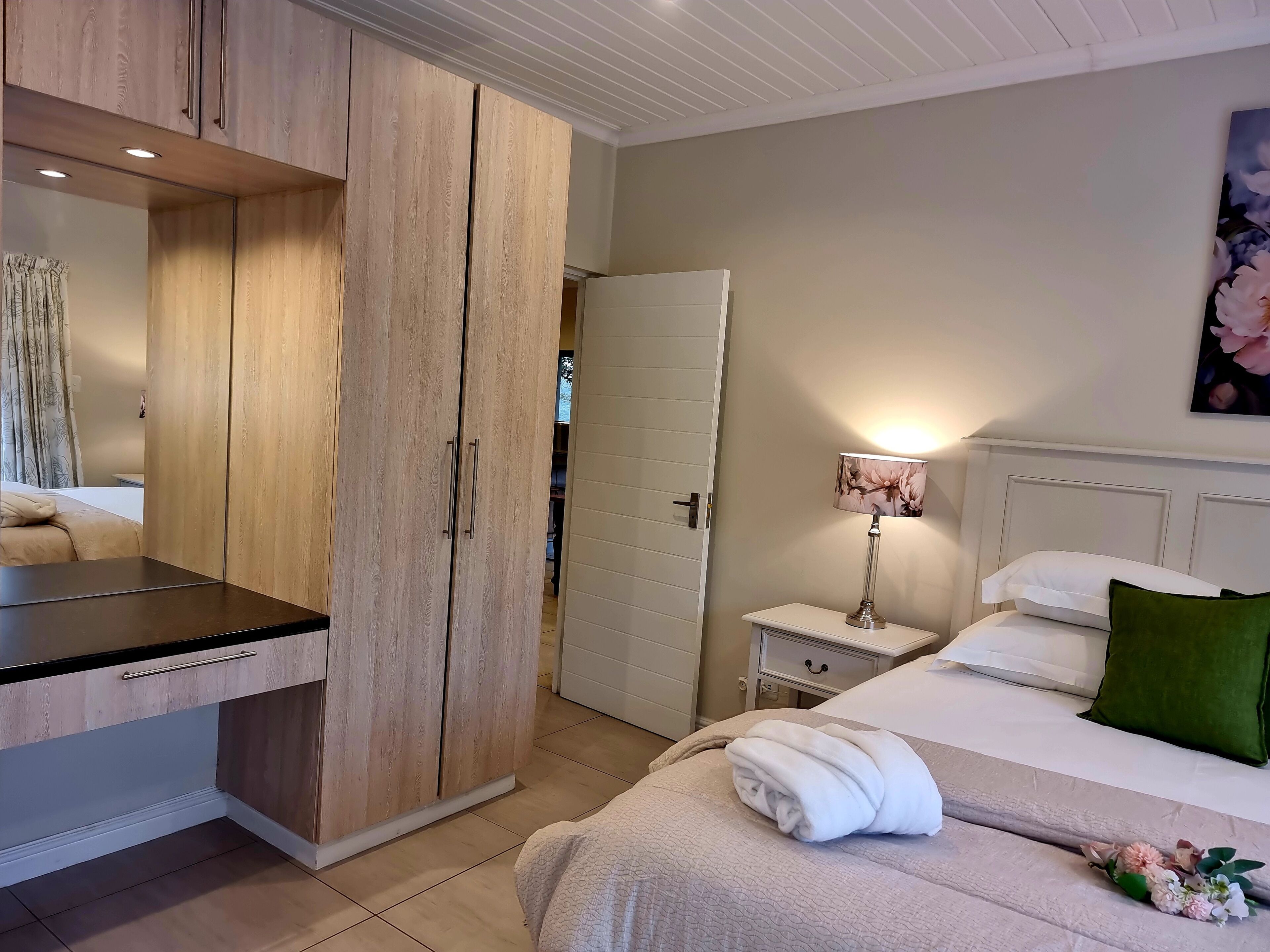 Un dormitorio con un armario de madera, una mesita de noche con lámpara, una cama con almohadas y una obra de arte floral en la pared.