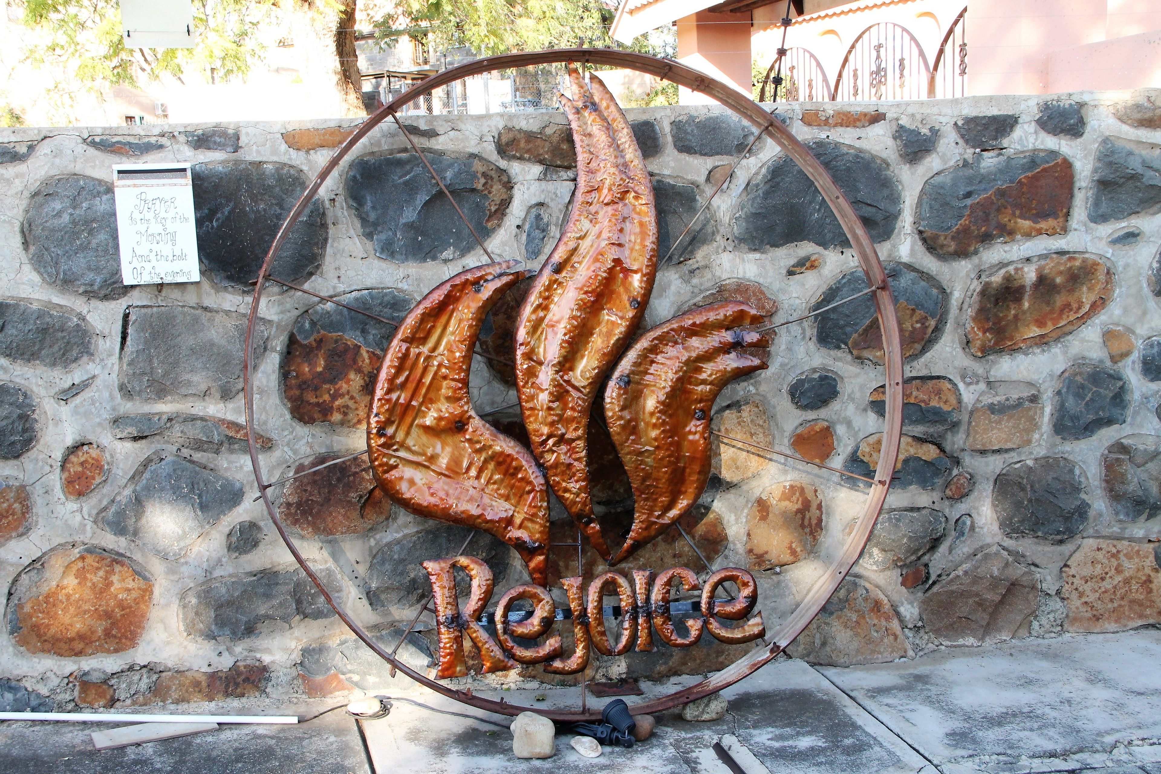 A stone wall with a metal sign featuring grilled fish and the word "Rejoice".
