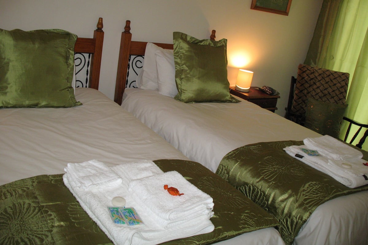 Habitación de hotel con dos camas, cada una con ropa de cama verde y blanca, y una mesita de noche con lámpara y control remoto.